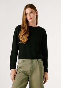 Melnā adīta džemperis ar garām piedurknēm, apaļu kakla izgriezumu, ribotām aprocēm un apakšmalu, kombīnēts ar gaiši olīvu zaļām biksēm. Gluda tekstūra, brīva piegriezuma stils.