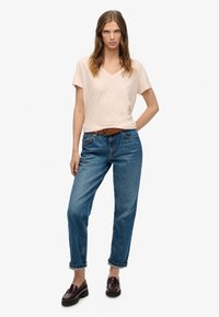 T-shirt rosa chiaro con scollo a V abbinata a jeans di denim blu larghi, risvolti arrotolati e cintura in pelle marrone. Le scarpe mocassino scure completano il look.