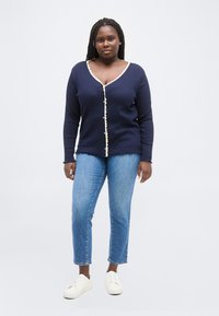 Γυναίκα με πλεξούδες, ντυμένη με μπλε σκούρο ζακέτα με λευκή λεπτομέρεια, μπλε τζιν και λευκά αθλητικά παπούτσια, στέκεται μπροστά σε ένα απλό φόντο.