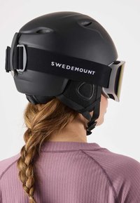 Czarny kask narciarski z otworami wentylacyjnymi, regulowanymi paskami i logo SWEDEMOUNT na tylnej części. Noszony z lustrzanymi goglami narciarskimi.