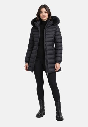 Donna che indossa un lungo cappotto invernale trapuntato nero con cappuccio rifinito in pelliccia, leggings neri e stivaletti neri, in piedi su sfondo bianco uniforme.