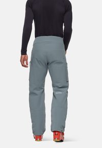 Pantalons de ski gris avec une taille haute, poches latérales zippées, genoux renforcés et poignets élastiques, associés à des bottes de ski orange.