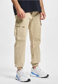 Man die beige cargobroek met elastische boorden en witte sneakers met blauwe accenten draagt, staand tegen een effen achtergrond.