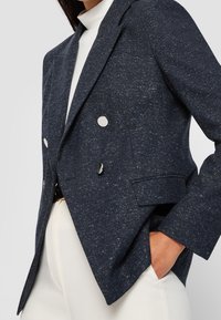 Mörkblå dubbelknäppt kavaj i en texturerad tyg med silverknappar och skräddarsydd passform, bärs över en vit höghalset topp.