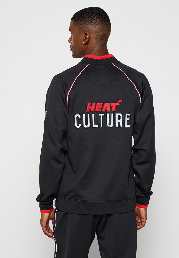 Uomo che indossa una giacca nera con rifiniture rosse e bianche, con "HEAT CULTURE" ricamato sul retro, in piedi contro uno sfondo chiaro e uniforme.