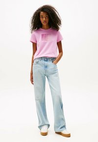 Camiseta de algodón de color rosa claro con logo en la parte frontal, combinada con jeans de pierna amplia de color azul claro. Zapatillas blancas con suelas marrones completan el atuendo.