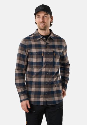 Mand iført en blå og beige ternet skjorte med brystlommer og sorte bukser, smilende og med blikket let til højre.