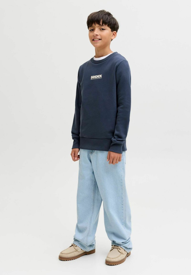 Ragazzo sorridente, indossa una felpa blu navy con la scritta "Bangkok", jeans azzurri e scarpe beige, in piedi contro uno sfondo chiaro e neutro.
