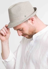 Világosszürke szőtt fedora kalap lapos koronával, széles peremmel és vékony szürke szalaggal. A modellen fehér gombos ing van, és a karimát lefelé tartja.