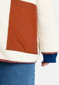 Sherpa fleece jack met een grote roestkleurige nylon patch. Kenmerken zijn blauwe gebreide manchetten, een gestructureerd witte buitenkant en een zachte binnenvoering.