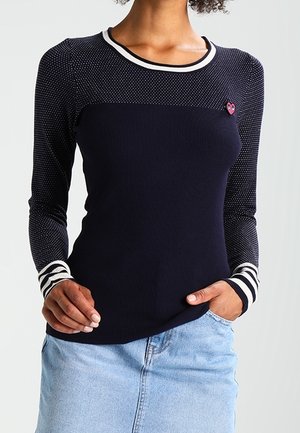 Frau trägt ein marineblaues Langarmshirt mit weißen gepunkteten Ärmeln, gestreiften Bündchen und Kragen, Herz-förmiger Anstecknadel, kombiniert mit einem hellblauen Jeansrock.