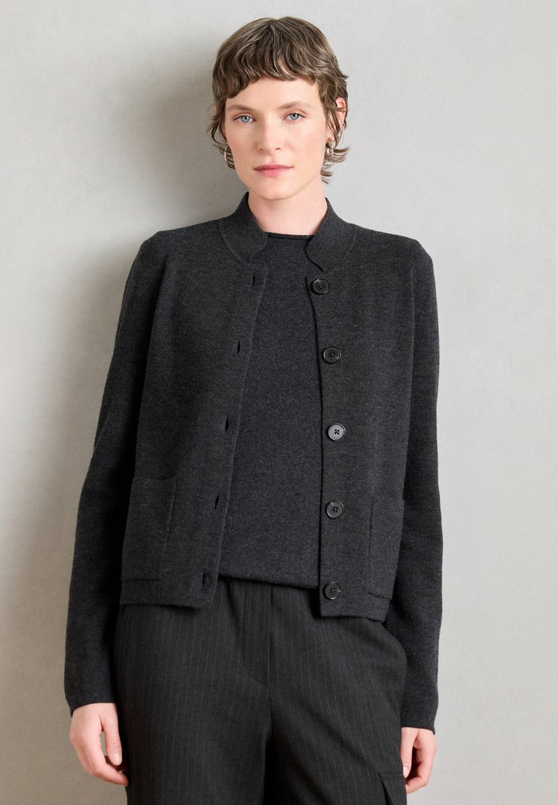 Cardigan gris oscuro hecho de una tela suave, con cuello redondo, cierre de botones y dos bolsillos. Llevado sobre una blusa a juego.