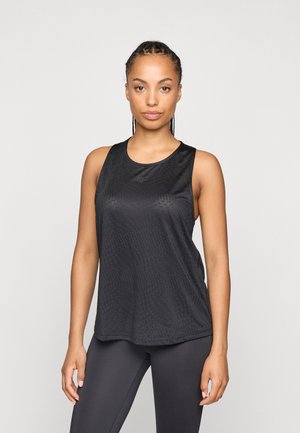 Jonge vrouw met gevlochten haar, gekleed in een zwarte mouwloze sporttop en zwarte legging, staand tegen een effen lichte achtergrond.