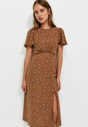 Mujer que lleva un vestido midi marrón con estampado floral, mangas cortas con volantes y una abertura frontal con detalles de encaje, de pie frente a un fondo blanco.