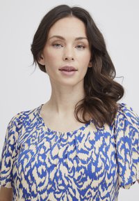 Kortärmad blus med ett blått och beige abstrakt mönster, rund halsringning och lätta veck framtill. Mjuk tygkänsla.
