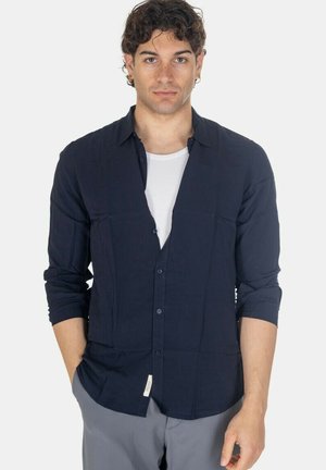 Giovane uomo con capelli ricci, che indossa una camicia blu scuro abbottonata sopra una canottiera bianca, mani in tasca, davanti a uno sfondo neutro.