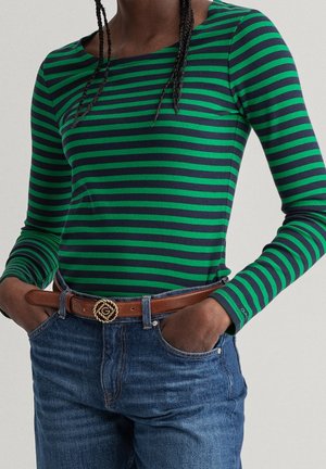Persoon draagt een groen en marineblauw gestreept shirt met lange mouwen, in een blauwe spijkerbroek gestopt met een bruine riem, handen in de zakken.