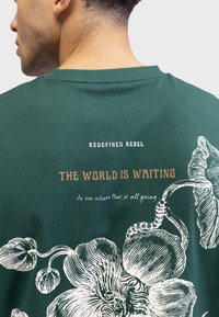 Grünes Baumwoll-T-Shirt mit einem großen weißen Blumenmuster und Text. Auf der Rückseite stehen die Phrasen "REDEFINED REBEL" und "THE WORLD IS WAITING."