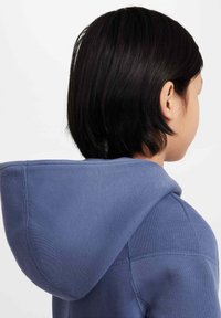 Pull à capuche bleu avec une grande capuche arrondie, des poignets côtelés et des coutures surpiquées. Texture douce et design décontracté, recouvrant partiellement les cheveux et le cou.