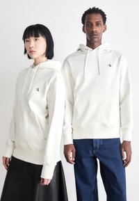 Felpe bianche in cotone con tasca frontale, dotate di un piccolo logo nero. Indossate da due modelli abbinate a una gonna di pelle e jeans blu.