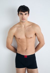 Schwarze Boxershorts mit einem roten Hugo-Label am Bund, in einer taillierten Passform und glatter Textur, getragen von einem männlichen Modell vor einem hellen Hintergrund.
