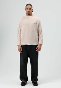 Beige fleece trui met lange mouwen, ronde hals en zwarte logodetails. Gecombineerd met losse zwarte denim broek en zwarte schoenen.