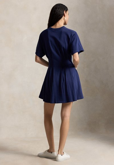 Frau steht mit dem Rücken zur Kamera, trägt ein marineblaues kurzärmeliges Kleid und weiße Turnschuhe vor einem neutralen Hintergrund.