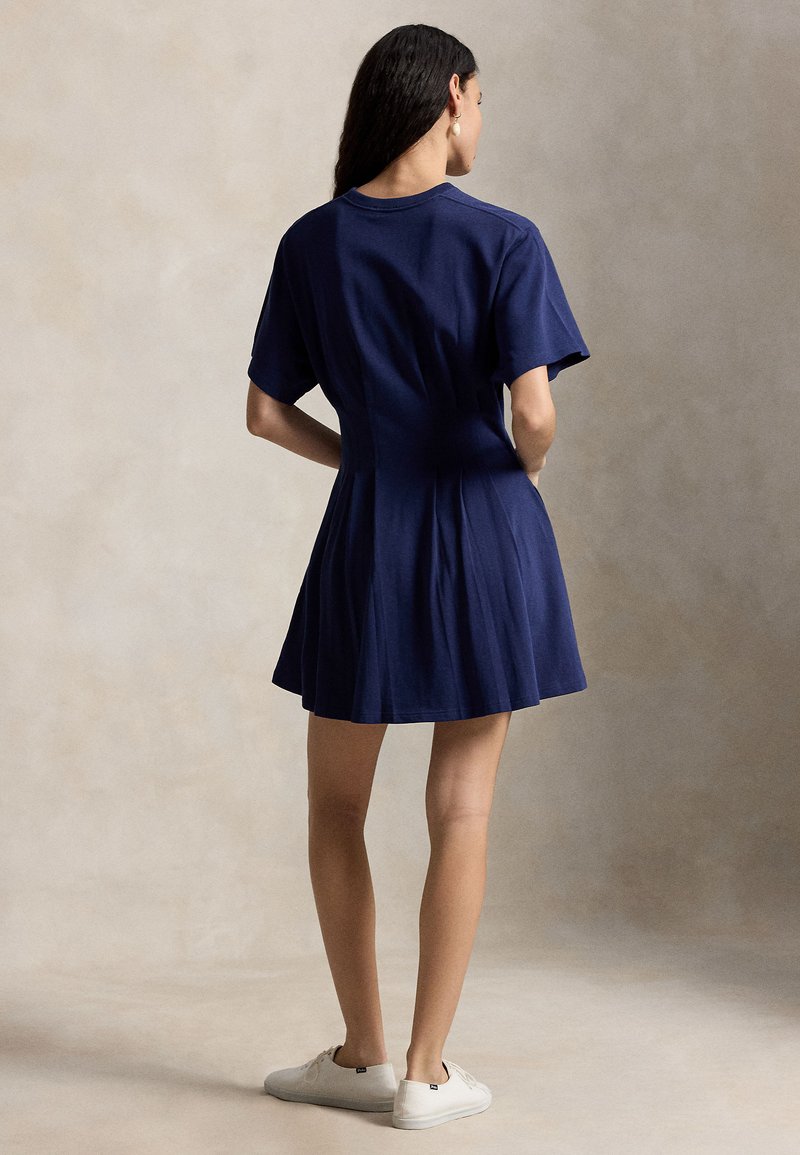 Frau steht mit dem Rücken zur Kamera, trägt ein marineblaues kurzärmeliges Kleid und weiße Turnschuhe vor einem neutralen Hintergrund.