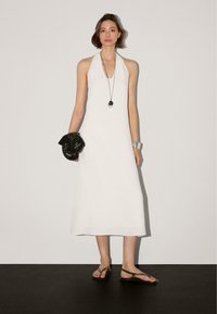 Robe halter en lin blanc avec une silhouette droite, sac tissé noir, bracelet en argent et sandales marron, sur un fond neutre.