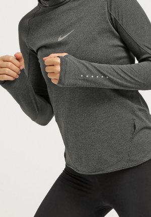 Persoon die een donkergrijze Nike sporttop met lange mouwen en duimgaten en zwarte legging draagt, geposeerd alsof ze aan het joggen of hardlopen is.