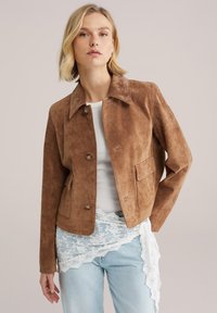 Bruine suède jas met een spitse kraag, knoopsluiting aan de voorkant en twee opgestikte zakken, gecombineerd met een licht tanktopje en versleten jeans.