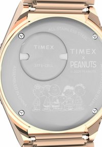 Urets bag i rustfrit stål har et cirkulært batteriområde, indgraveret med "TIMEX x PEANUTS" og karakteromrids. Gyldne accenter.