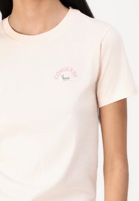 Ljusrosa bomullströja med rund halsringning. Har en liten grön och rosa grafik av en myra och ordet "CONVERSE" ovanför.