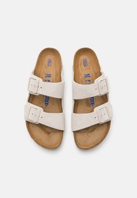 Sandales en suède blanc avec deux brides ajustables, semelle en liège plate et doublure en cuir lisse. Détails de boucle sur chaque bride.