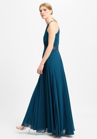 Türkisfarbenes ärmelloses Kleid mit tailliertem Oberteil und fließendem Rock. Detaillierte Spitzenakzentuierung an der Taille und subtile Falten für Bewegungsfreiheit.