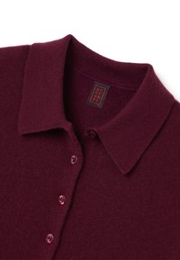 Maglione a maglia bordeaux con colletto, caratterizzato da cinque bottoni rosa lungo la patta e un'etichetta di stoffa con testo rosso e nero.