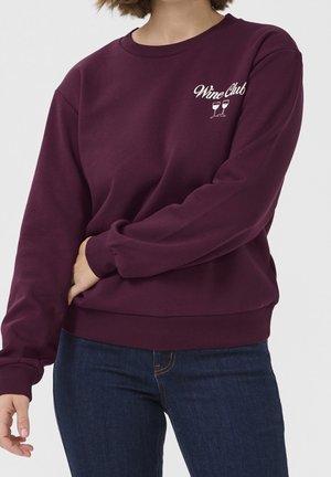 Nainen, joka pukeutunut viininpunaisen sävyiseen collegepaitaan, jossa rintakehällä "Wine Club" -teksti ja kaksi viinilasin kuvaa, yhdistettynä tummansinisiin farkkuihin.