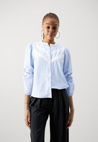 Light blue striped blouse with lace detailing, button front, puffed sleeves, and a rounded neckline, paired with black trousers.