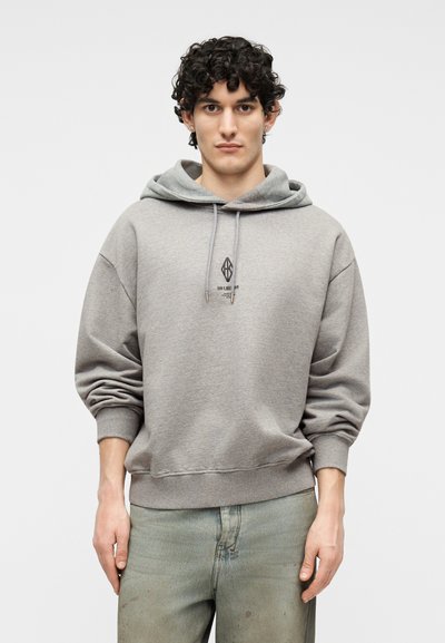 Νεαρός άνδρας με σκουρόχρωμα σγουρά μαλλιά, φορώντας γκρι φούτερ με κουκούλα με ένα μικρό μαύρο λογότυπο και ανοιχτόχρωμα ξεθωριασμένα τζιν, που στέκεται μπροστά σε ένα απλό φόντο.