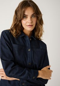 Frau mit schulterlangen, hellbraunen Haaren, die eine dunkelblaue Jeansjacke trägt, die Arme verschränkt hat und sanft in die Kamera schaut, vor einem schlichten Hintergrund.