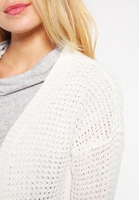 Frau mit blonden Haaren, die ein graues Oberteil und eine weiße Strickjacke trägt, von Hals bis Schulter vor einem weißen Hintergrund gezeigt.
