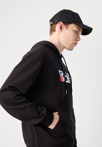 Svart hettegenser med kengurulomme, med et grafisk design i hvitt og rødt. Svart caps med mønster og justerbar rem.