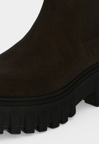 Gros plan d'une botte Chelsea en daim brun foncé avec une épaisse semelle noire à crampons et un panneau latéral élastique.