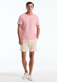 T-shirt rose à rayures avec un col rond, associé à un short beige. Modèle portant des baskets blanches se tient devant un fond uni.