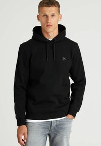 Sudadera negra de tela suave con un bolsillo frontal. Cuenta con una capucha con cordón y un pequeño logo en el pecho. Se lleva con jeans claros.
