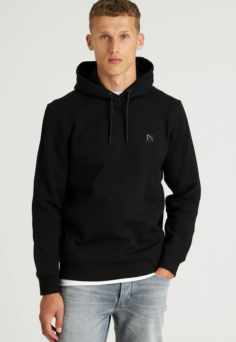 Sudadera negra de tela suave con un bolsillo frontal. Cuenta con una capucha con cordón y un pequeño logo en el pecho. Se lleva con jeans claros.