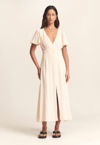 Crèmekleurige jurk tot op de knie met korte pofmouwen, diepe V-hals, knoopsluiting aan de voorkant en een voorsplit. Gladde stoftextuur.