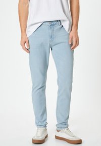Jeans de denim azul claro con un corte ajustado, diseño de cinco bolsillos y cierre delantero de botón. Llevados con zapatillas blancas y una camiseta blanca.