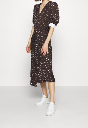 Kvinde iført en brun midi omslagskjole med hvide prikker, hvide manchetsærmer og hvide sneakers, stående mod en ensfarvet baggrund.