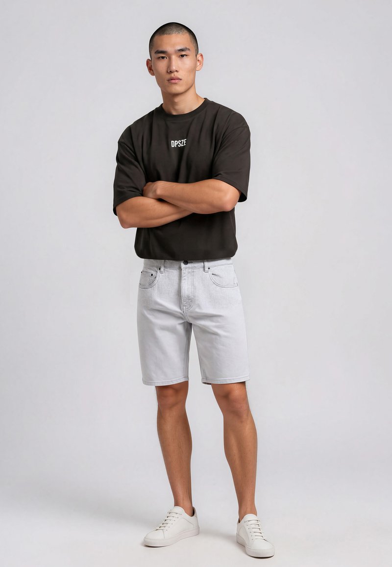 Homme debout, bras croisés, portant un t-shirt noir à motif graphique, un short en jean gris clair et des baskets blanches sur fond uni.